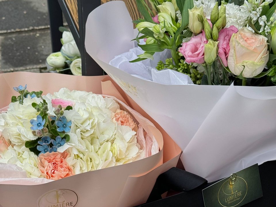 Zwei Arrangements in LUMIÈRE-Verpackung auf LUMIÈRE-Tragetaschen: weiße Hortensie mit blauen Akzenten in pfirsichfarbenem Papier und ein Strauß mit rosa Rosen in weißem Papier